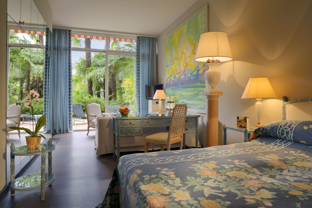 Sous la lumière du Tessin, la chambre s’ouvre sur un jardin luxuriant où palmiers et élégance italienne invitent à la rêverie.